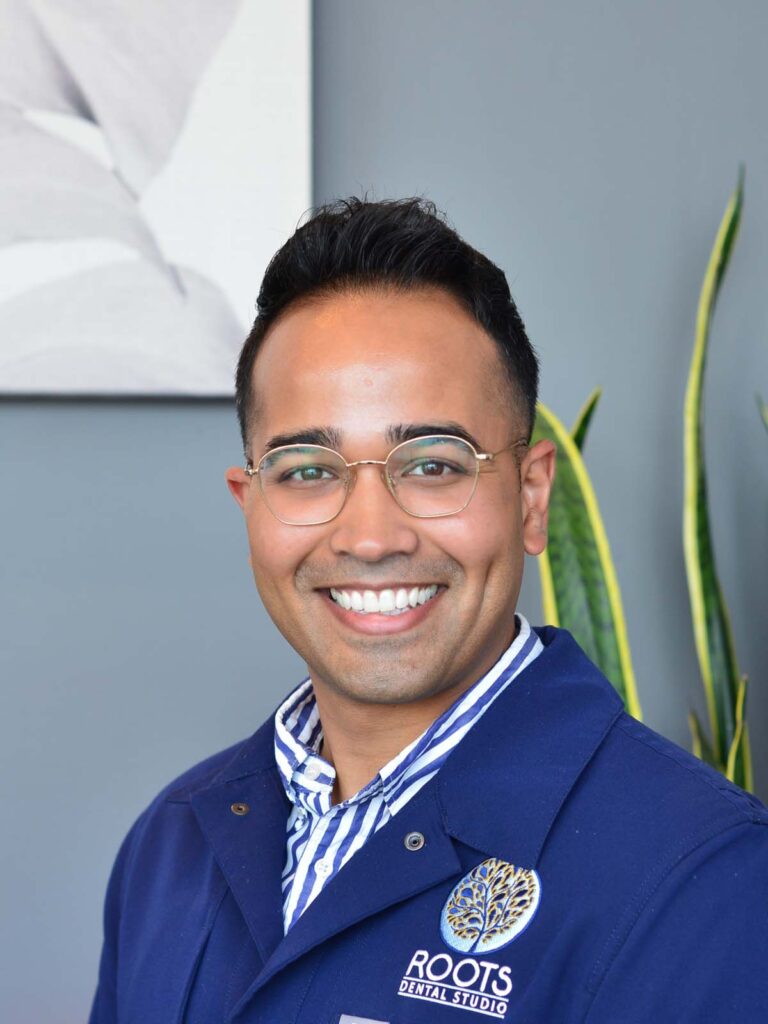 The image shows a man wearing an eyeglasses and a blue coat with the logo 'ROOTS DENTAL STUDIO' clearly visible on the left side.