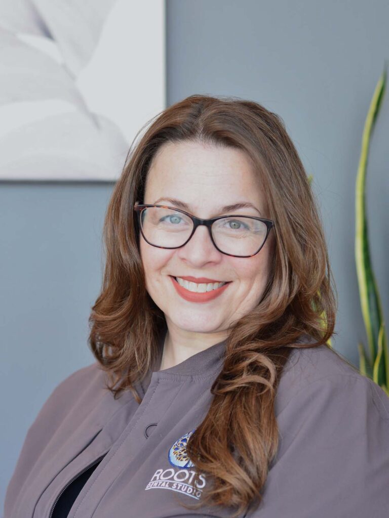 An image of a girl wearing a black eyeglasses with a medium length wavy brown hair in a semi side view pose. She is wearing a dark gray uniform with a logo, under of it is a text "ROOTS DENTAL STUDIO" that placed on the right side.