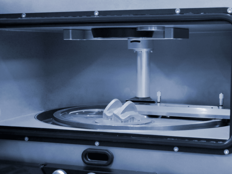 The image shows a 3D printer in operation, printing a complex-shaped part with visible support structures and mechanical components.