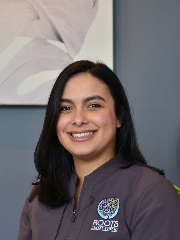 An image of a girl with black straight hair in a semi side view pose. She is wearing a dark gray uniform with a logo, under of it is a text "ROOTS DENTAL STUDIO" that placed on the right side.