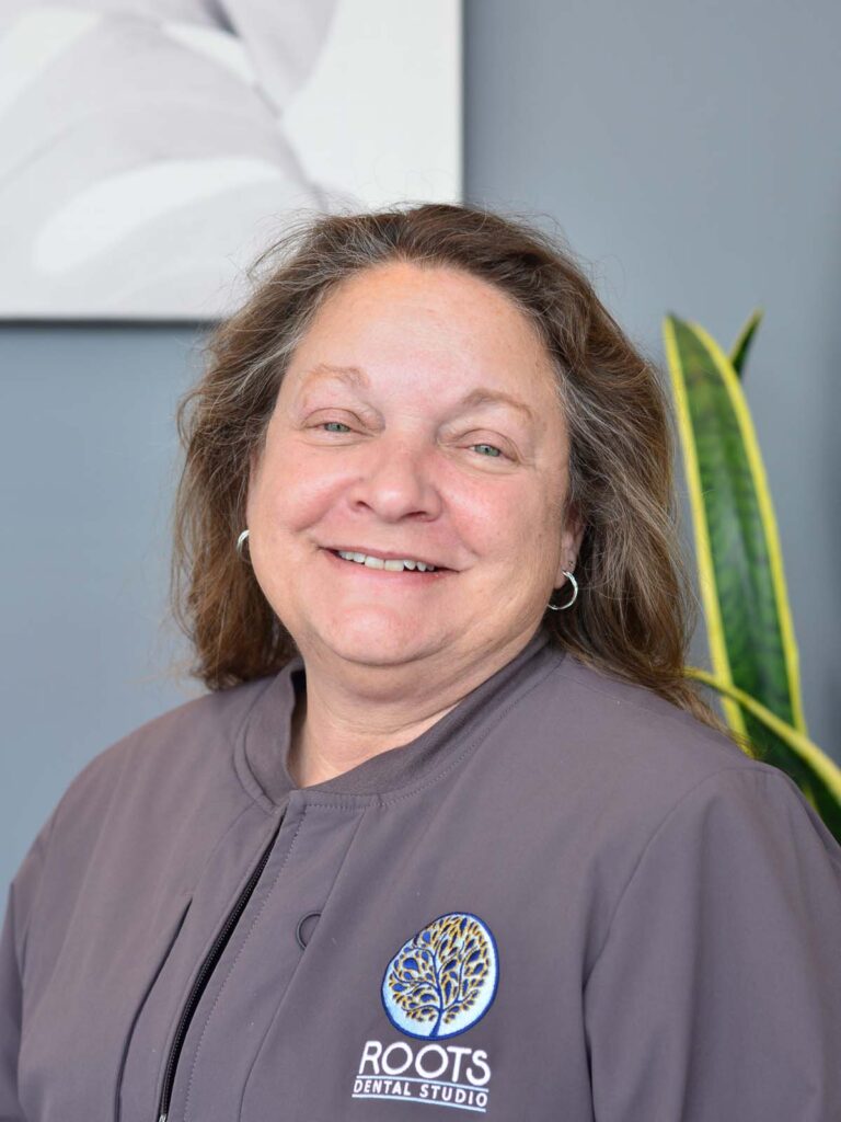 An image of a girl smiling in a semi side view pose. She is wearing a dark gray uniform with a logo, under of it is a text "ROOTS DENTAL STUDIO" that placed on the right side.