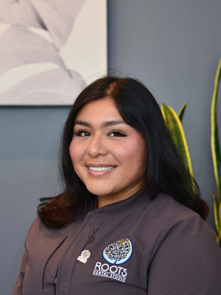An image of a girl smiling in a semi side view pose. She is wearing a dark gray uniform with a badge beside the logo, under of it is a text "ROOTS DENTAL STUDIO" that placed on the right side.