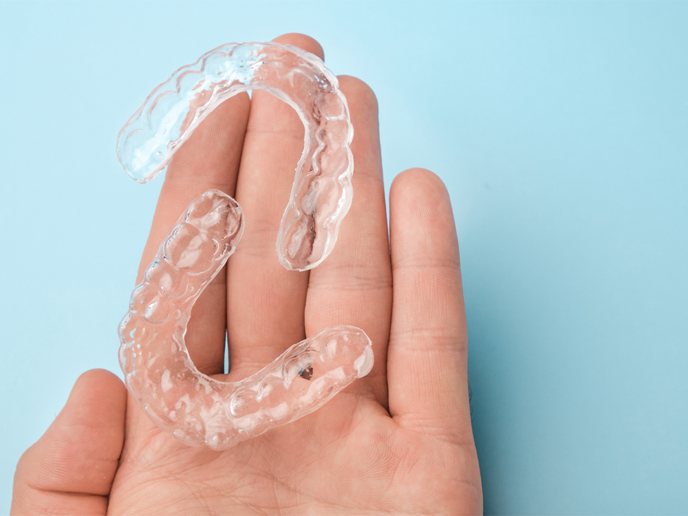 The image shows a palm with a pair of clear dental retainers on it. The background color is baby blue.