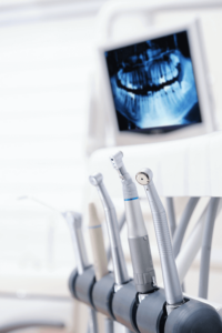The image shows a close-up view of dental equipment, specifically several dental handpieces, which are tools used by dentists for various procedures. In the background, there is a monitor displaying a dental X-ray image of a patient's teeth.