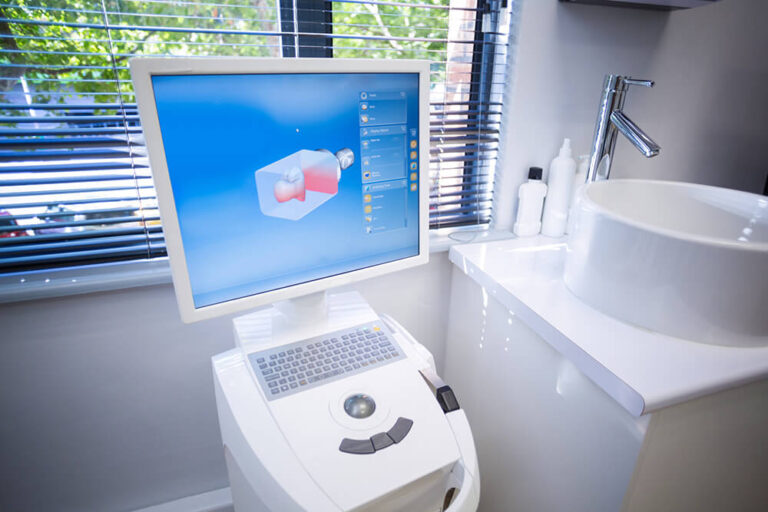 The image shows a dental CAD/CAM (Computer-Aided Design and Computer-Aided Manufacturing) system in a dental office. The system includes a monitor displaying a 3D model of a dental restoration, likely a crown or a bridge, and a keyboard with a trackball for navigation.