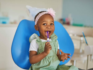 The image shows a young child sitting on a blue chair, in a dental office. The child is wearing a green polka-dotted dress with a white shirt underneath and a pink headband with a flower. The child is holding a toothbrush.