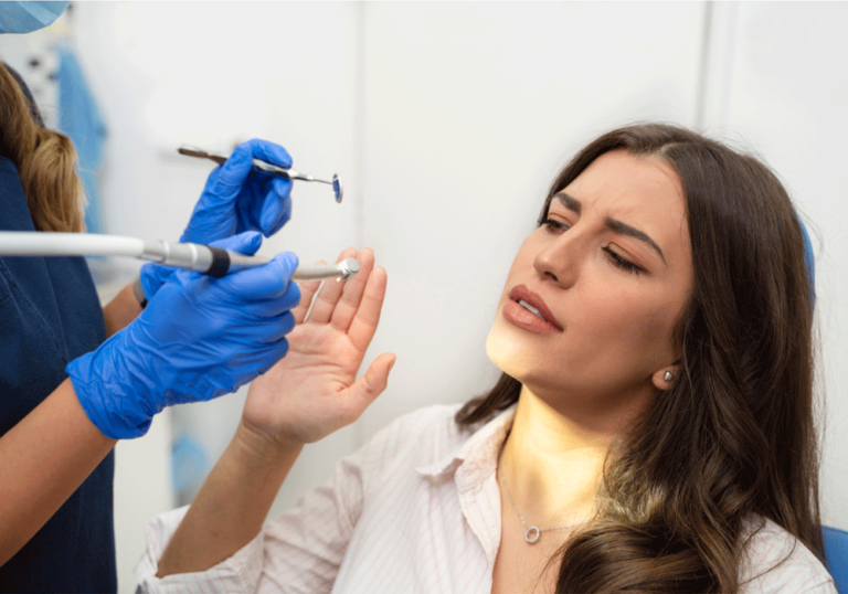 A dental professional wearing blue gloves is performing a dental procedure on a patient. The patient, appearing anxious, shows a hand sign while the dental professional examines their teeth using dental instruments.