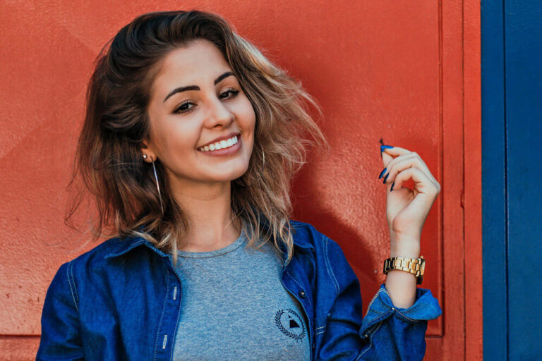 A girl smiling in a denim shirt over a grey t-shirt stands against a red background, holding a small object in one hand and wearing a gold wristwatch.